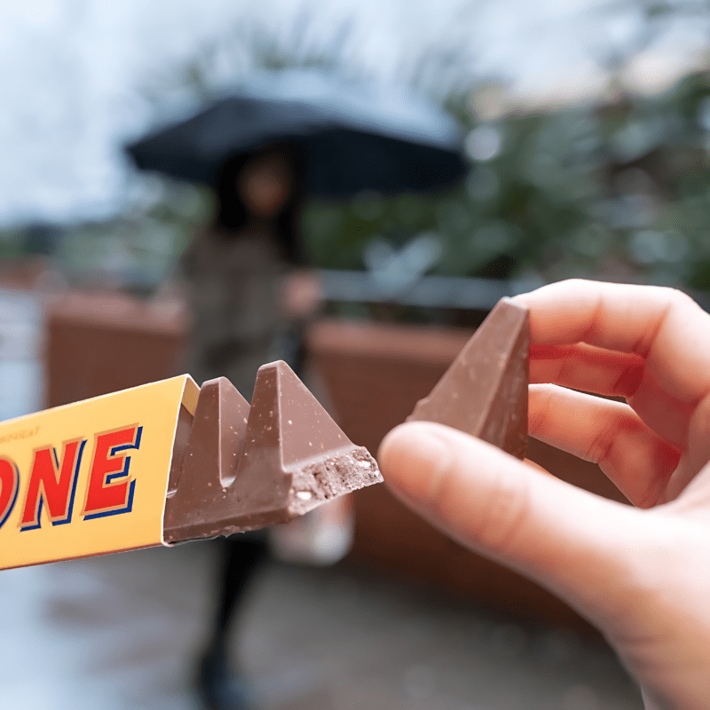 Top 10 Schweizer Schokoladenmarken 3 Erleben Sie den kultigen Geschmack von Schweizer Schokolade, wenn eine Hand nach einem Stück Toblerone greift, einem beliebten Klassiker unter den Schweizer Schokoladenmarken. Genießen Sie die cremige Textur und den ausgeprägten Honig-Mandel-Nougat-Geschmack dieses berühmten Meisterwerks der Süßwarenherstellung.