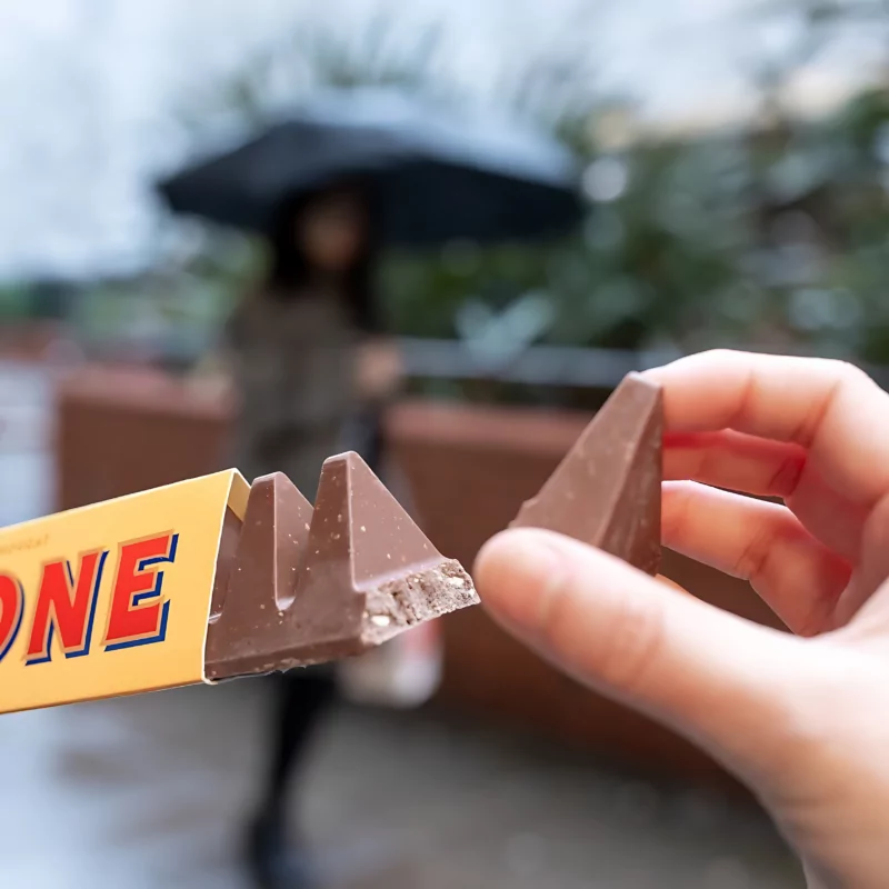 Experience the iconic taste of Swiss chocolate as a hand reaches for a piece of Toblerone, a beloved classic among top Swiss chocolate brands. Delight in the creamy texture and distinct honey-almond nougat flavor of this renowned confectionery masterpiece.